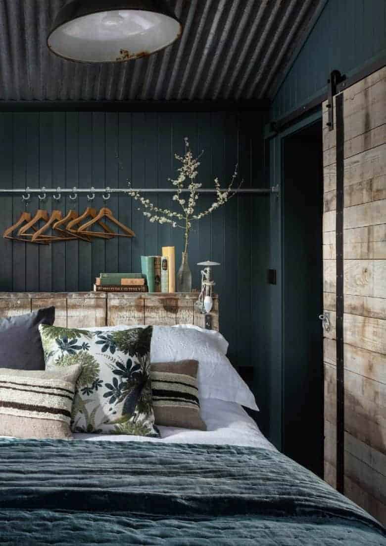 modern rustic bedroom decoration idea using reclaimed wood as a bedhead, industrial lighting, dark vintage fabrics, teal velvet, textural knitted cushions and crisp white linen bedding with dark teal tongue and groove walls #modern #rustic #bedroom #reclaimed #teal