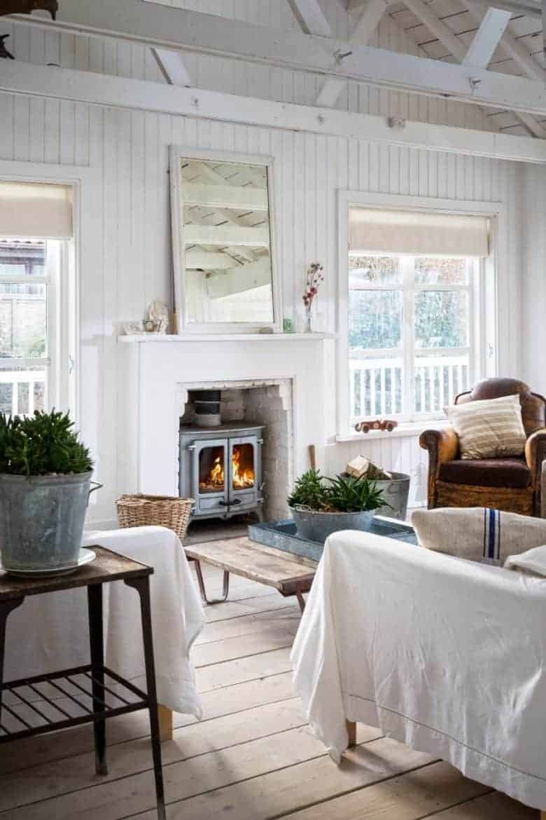 coastal cottage living room interior with white tongue and groove walls, rustic floor boards, white linen slip covers, vintage linen grainsack cushions, old leather armchair, wood burner white fireplace and old galvanised tubs with plants #white #cottage #livingroom