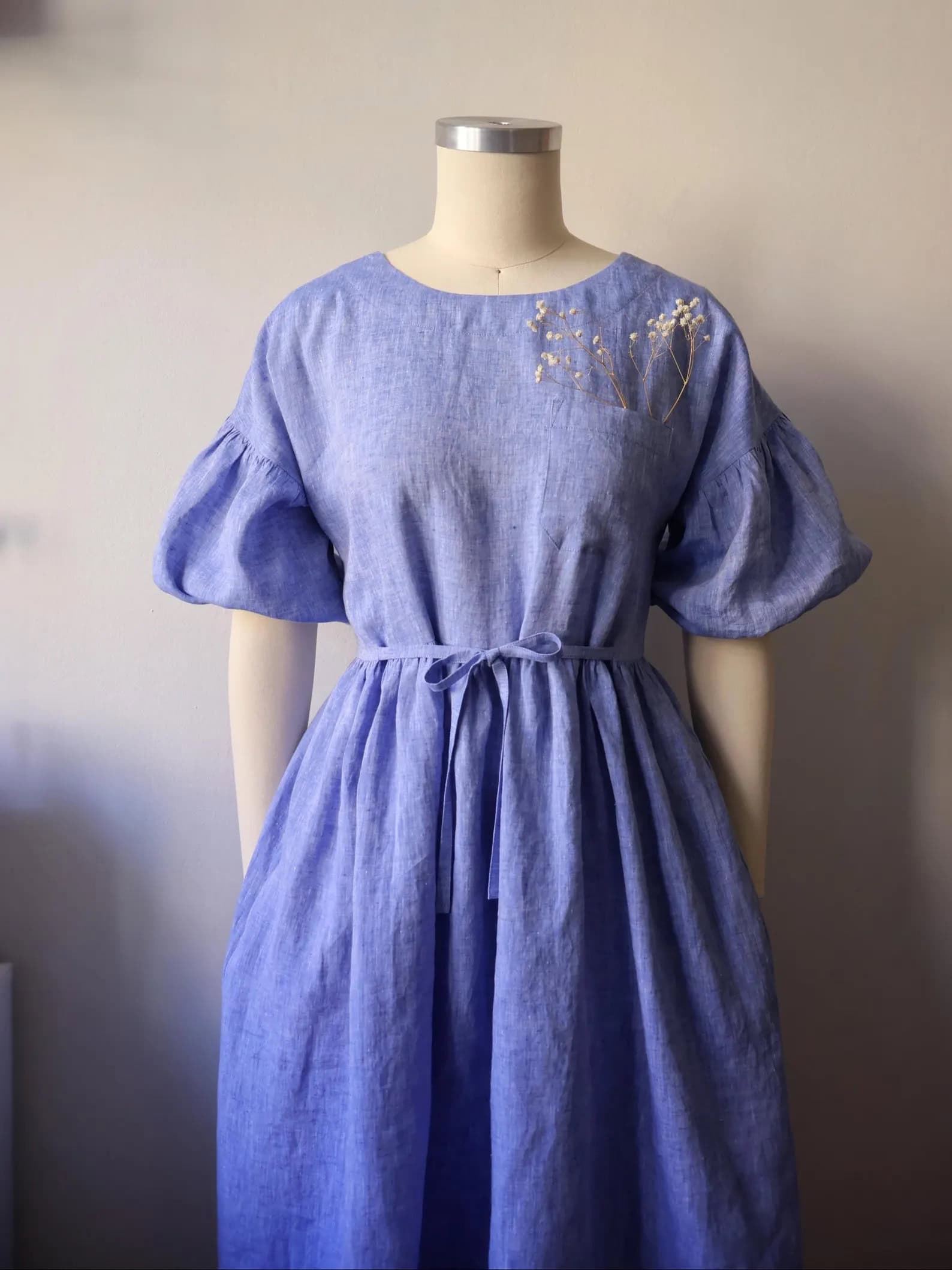 fern linen dress sewing pattern.relaxed silhouette, designed for comfort and everyday wear. It features a round neckline, a slightly dropped shoulder, and a gathered skirt that falls in midi length. The waistline is with self-fabric ties, offering adjustable shaping. The chest pocket and side seam pocket